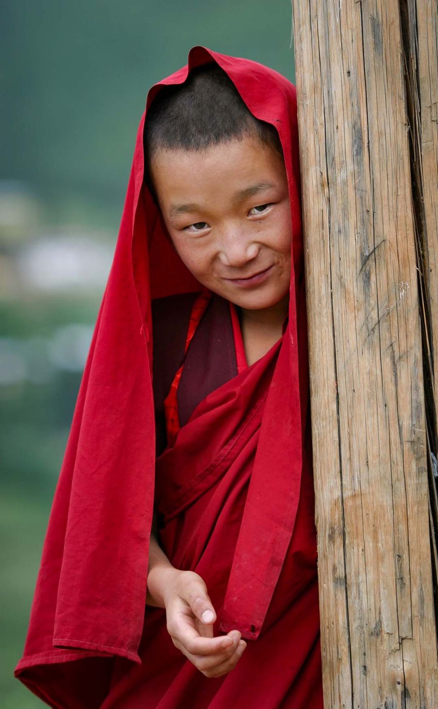 A young monk in crimson robes leaning around a temple doorpost