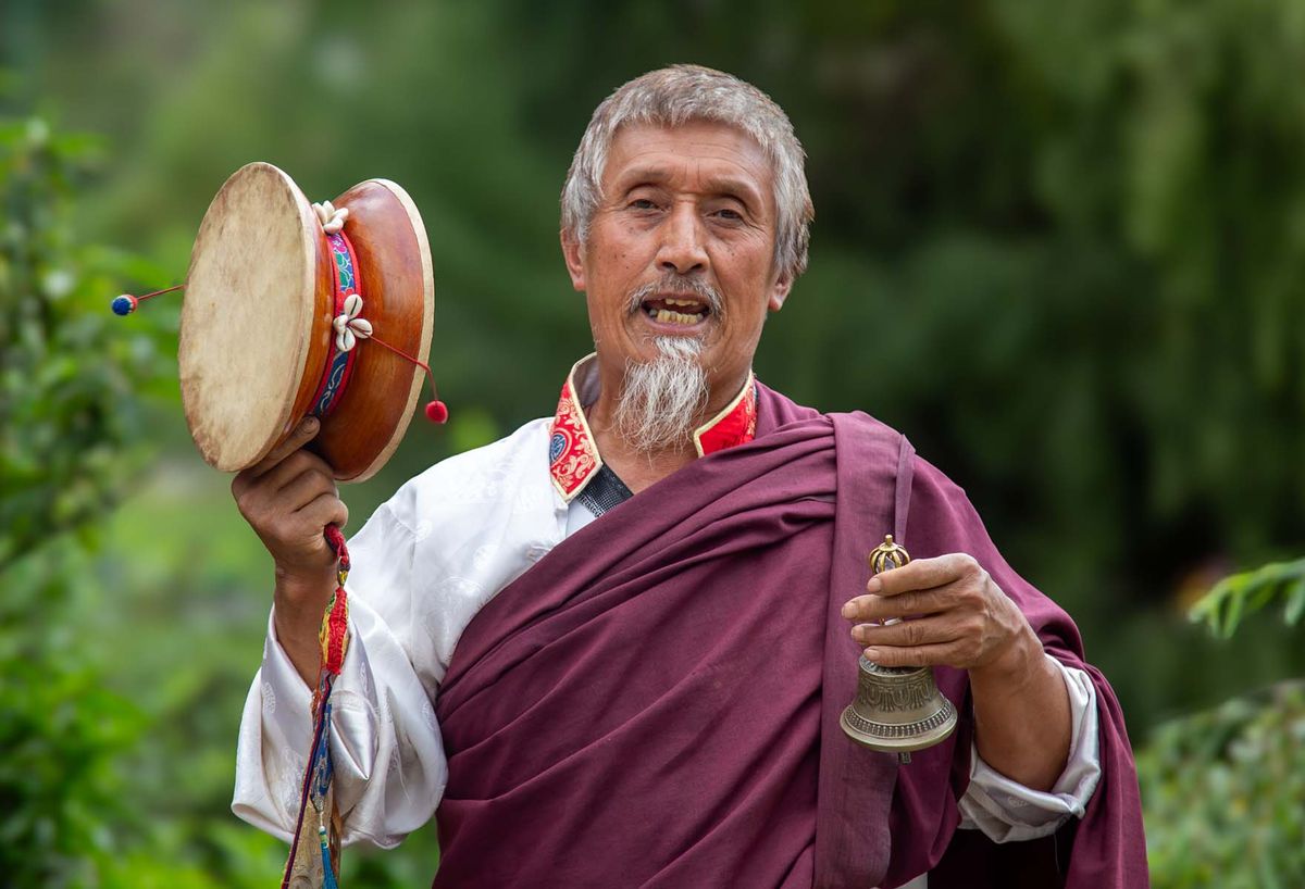 An elder in maroon robes holding a ritual hand drum and bell