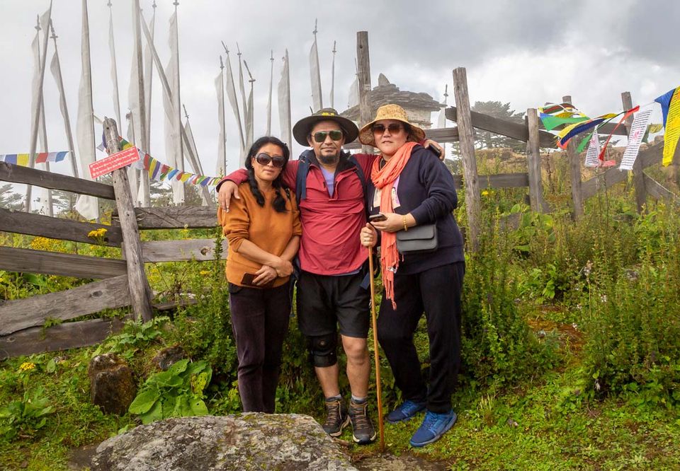 Three hikers pausing beside prayer flags on a green hillside