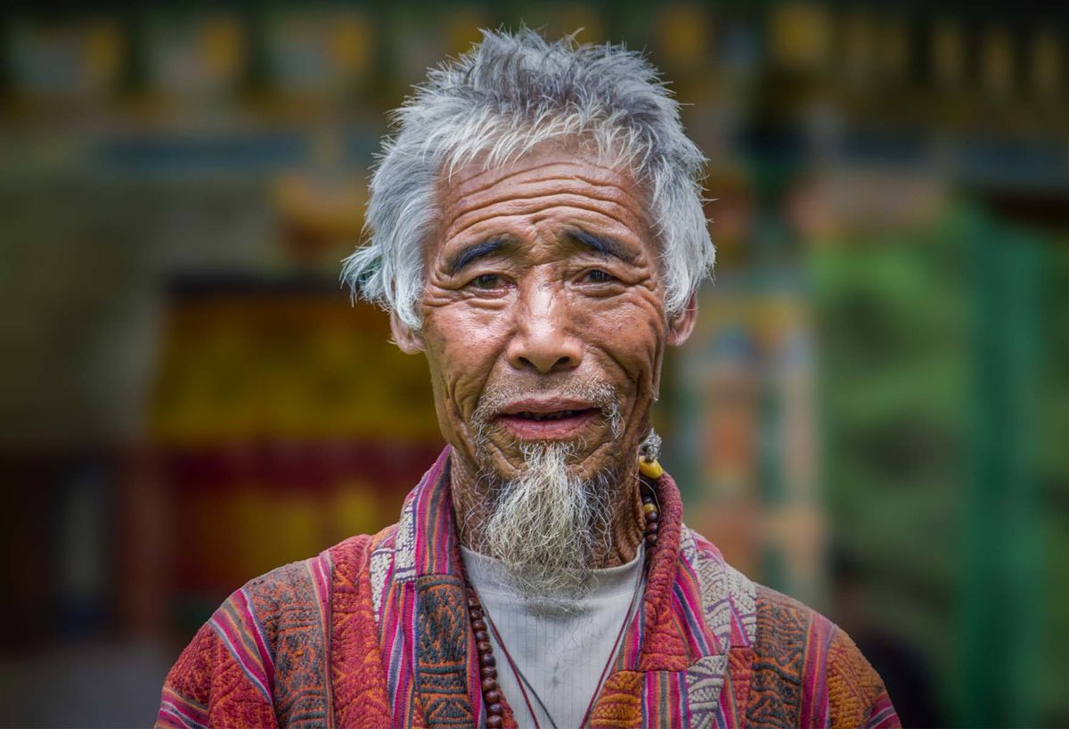 A bearded Bhutanese elder in a patterned robe looking to camera