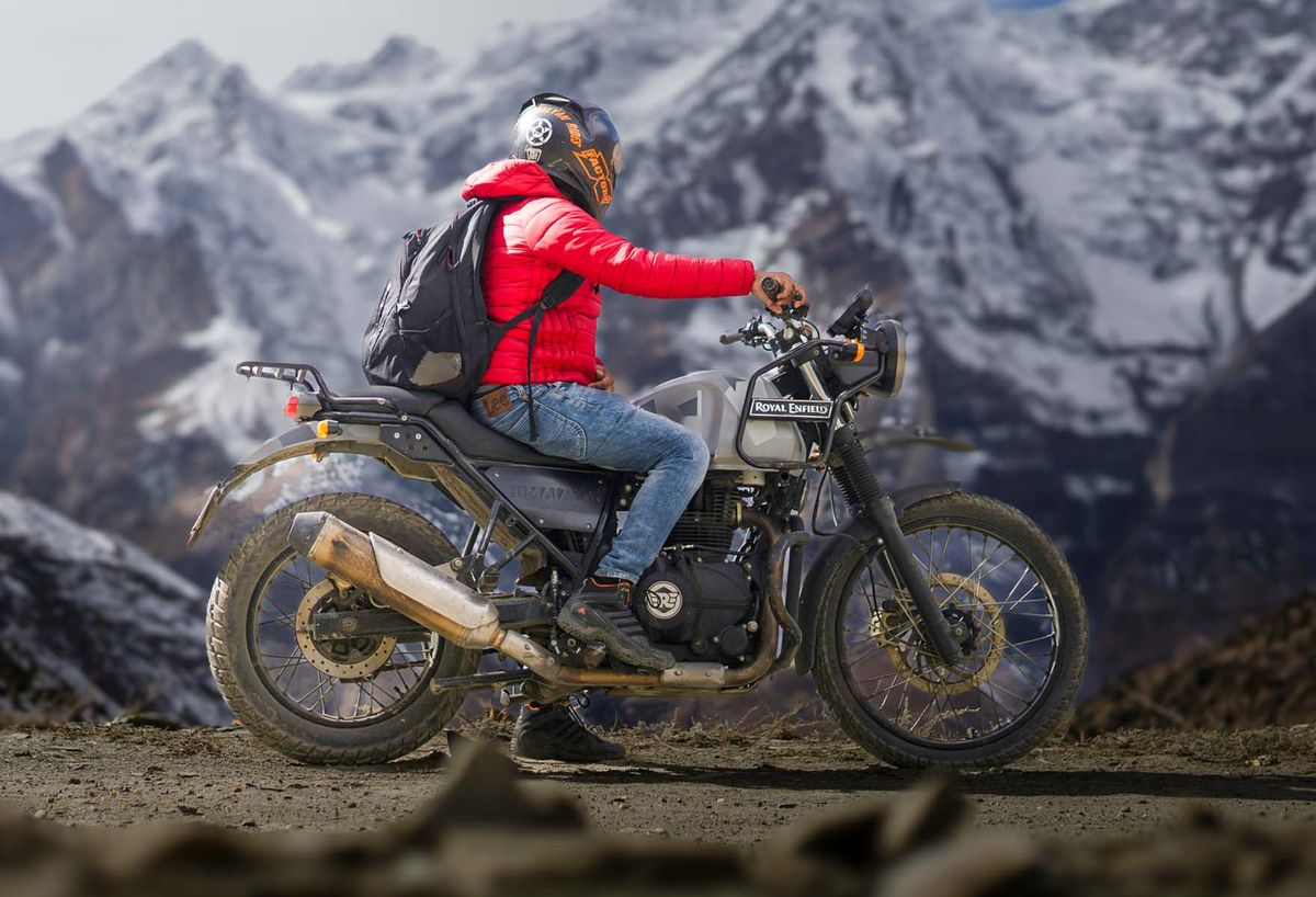 A rider in a red jacket on a mountain road below snowy peaks