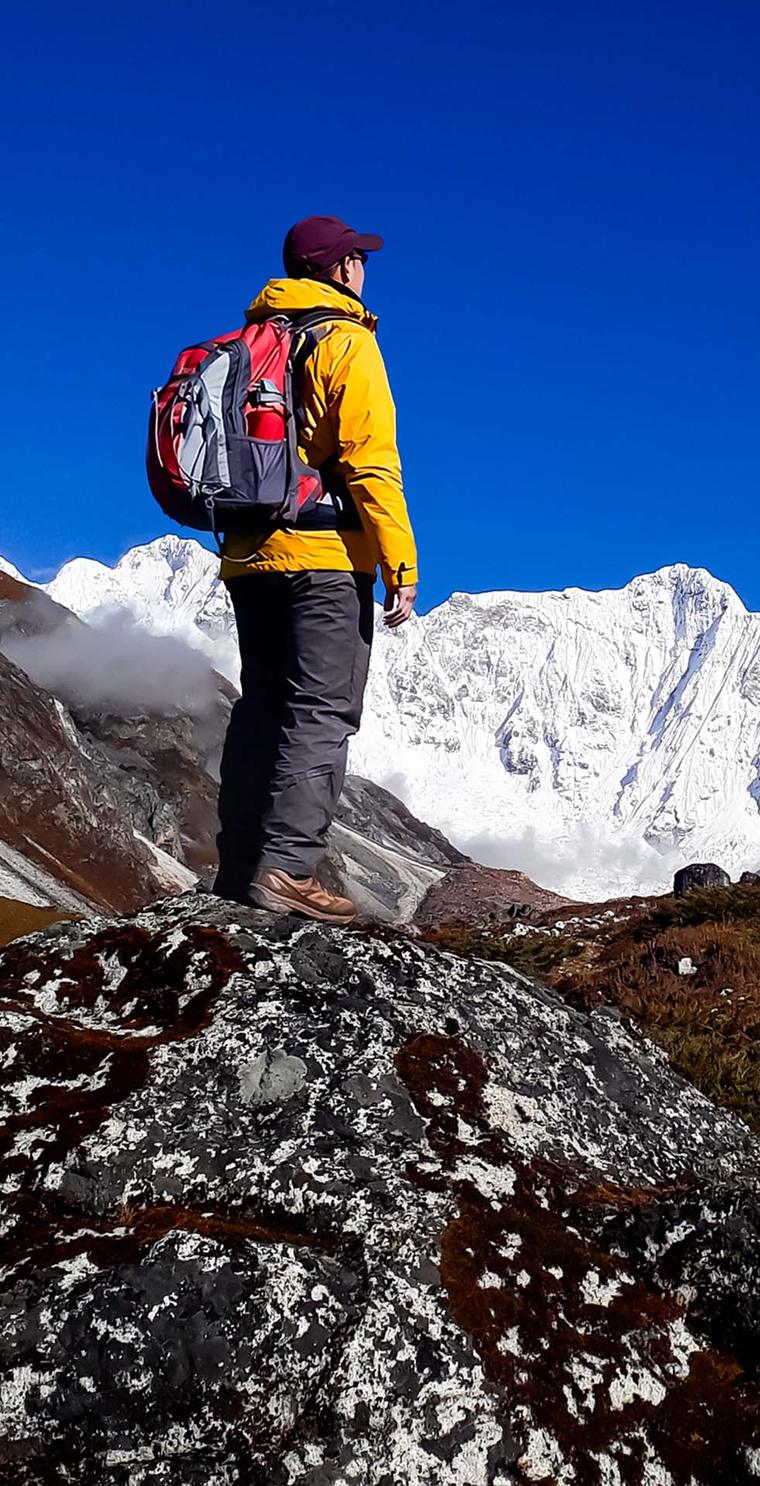 A trekker in a yellow jacket on a boulder facing snowy peaks