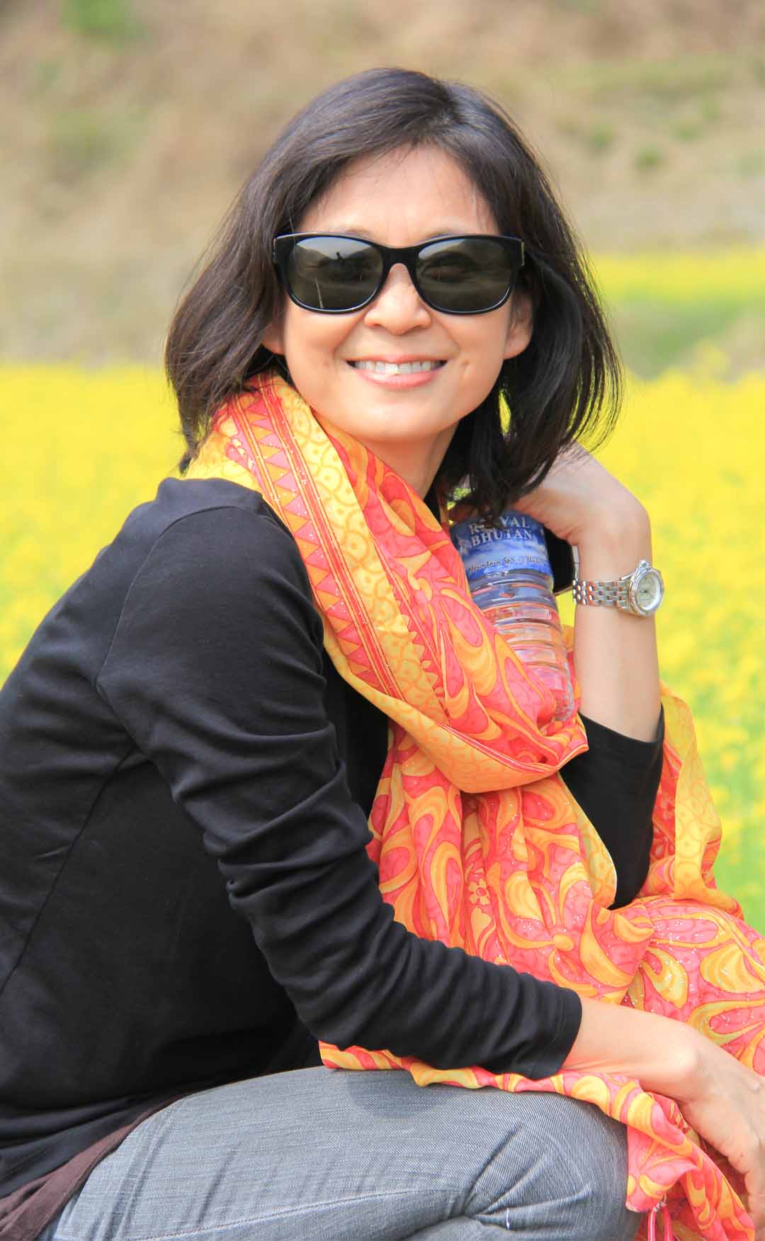 Hana Scoda in sunglasses and a bright orange scarf, seated in a field of yellow mustard flowers