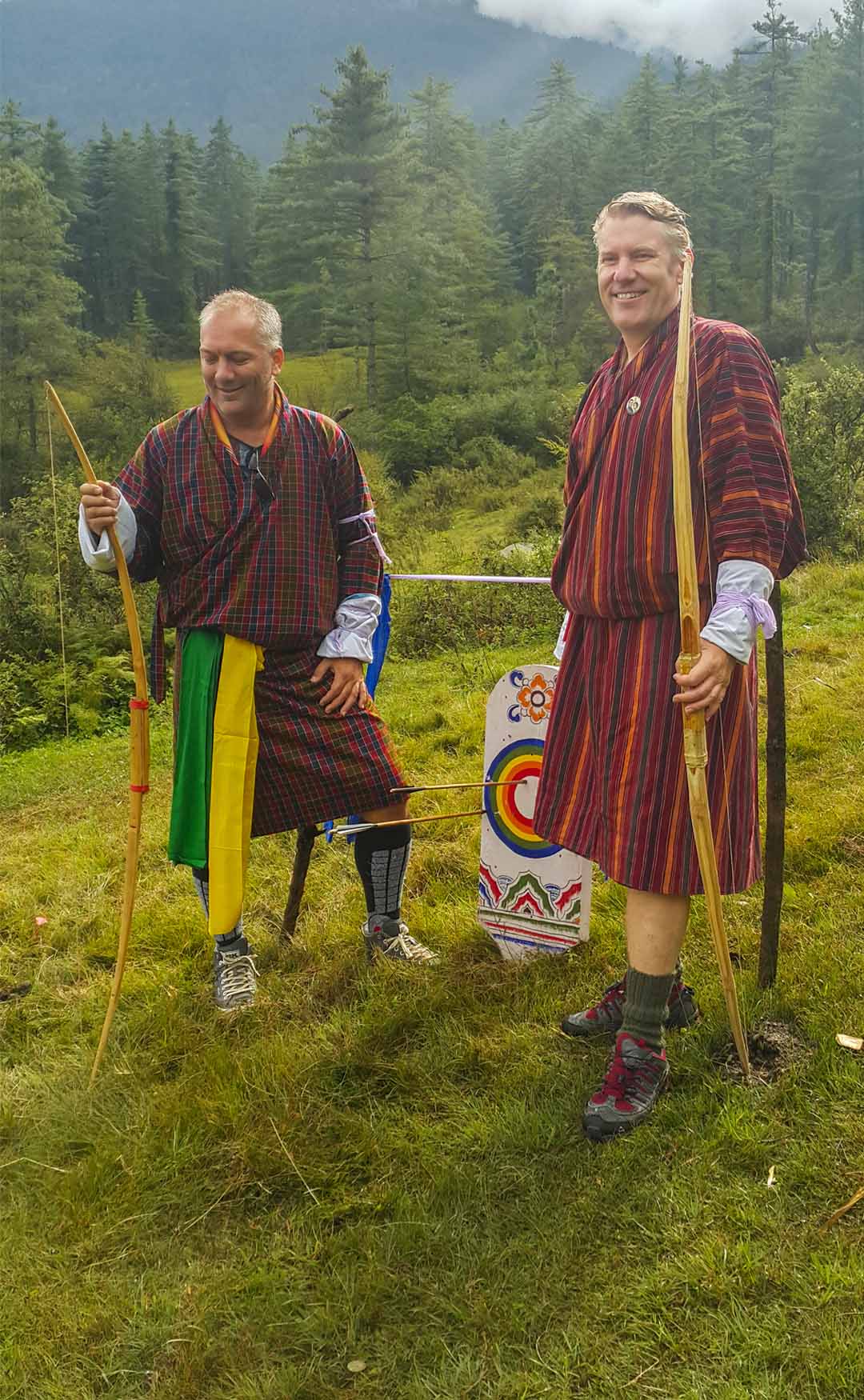 Markus and Michael in traditional Bhutanese gho, holding bamboo bows at an archery range in the forest